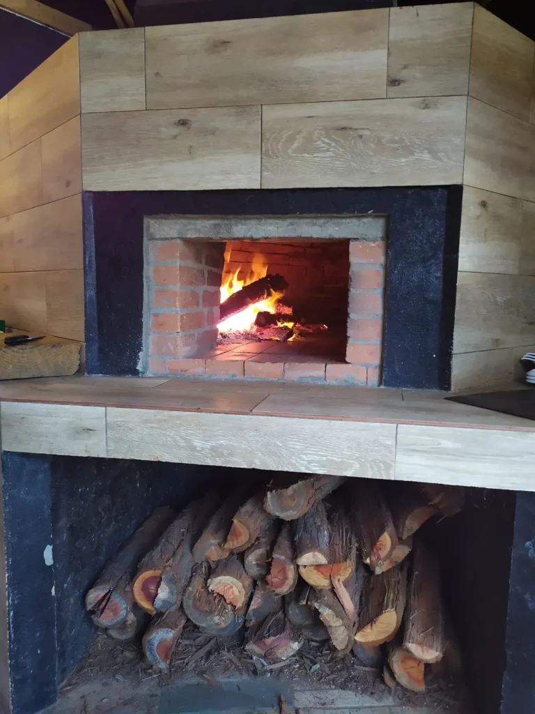 Four à bois allumé où cuisent les pizzas, bûches rangées dessous